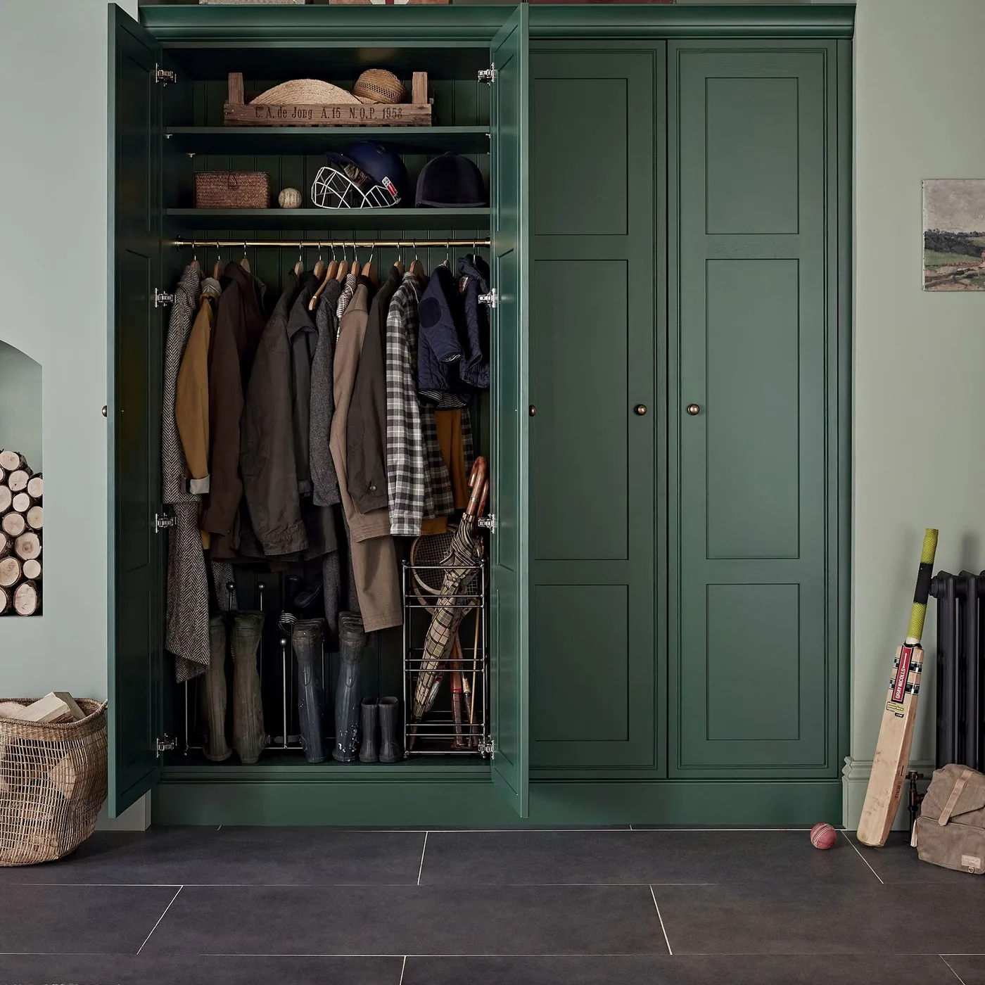 Deep green mudroom cupboard with one open door, hanging coats, upper hat shelves, boots and umbrella storage, beside a dark tiled floor.