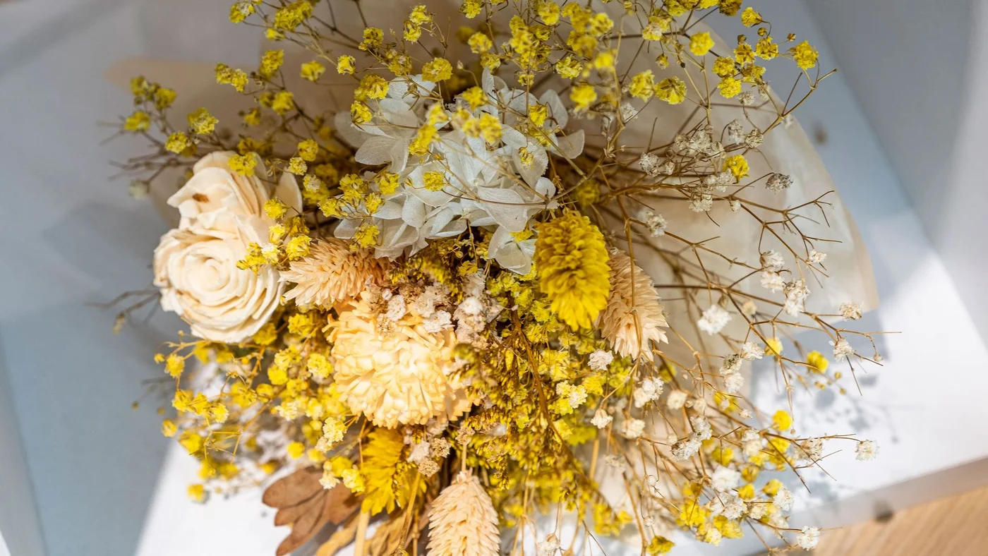 A close view of a dried bouquet shows yellow branching flowers, cream flower heads and pale grasses against light fabric.