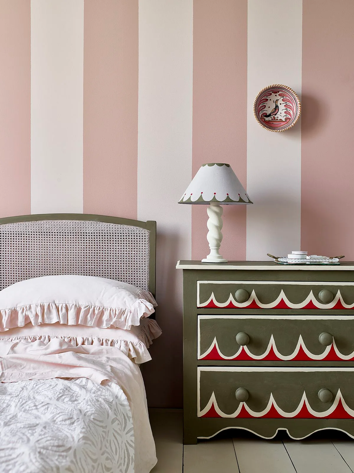 Broad pink-and-white wall stripes rise behind an olive headboard beside an olive cabinet with red-and-white scalloped drawer decoration