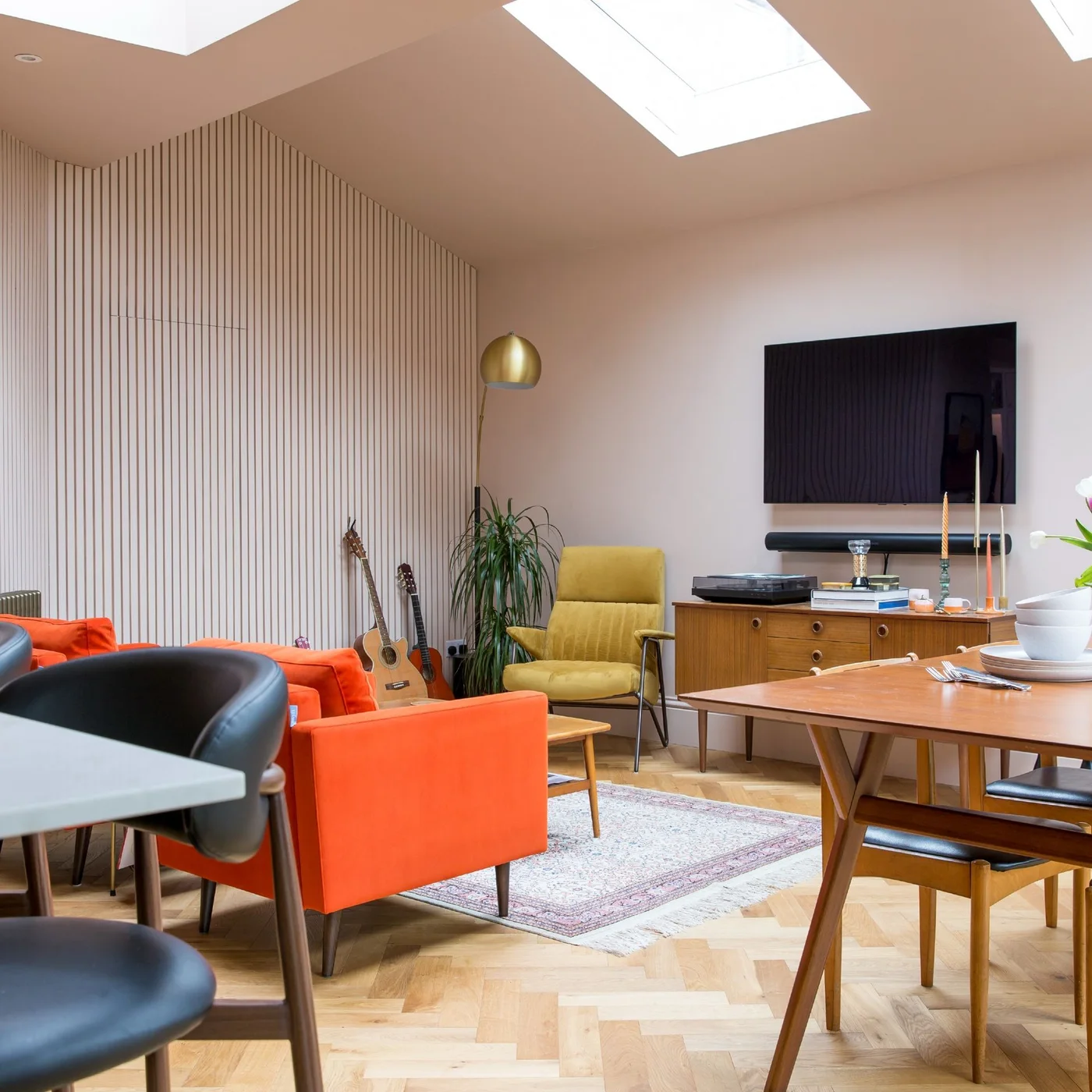 An orange sofa and yellow chair form a sitting area beside a dining table beneath roof glazing and a pale slatted wall.