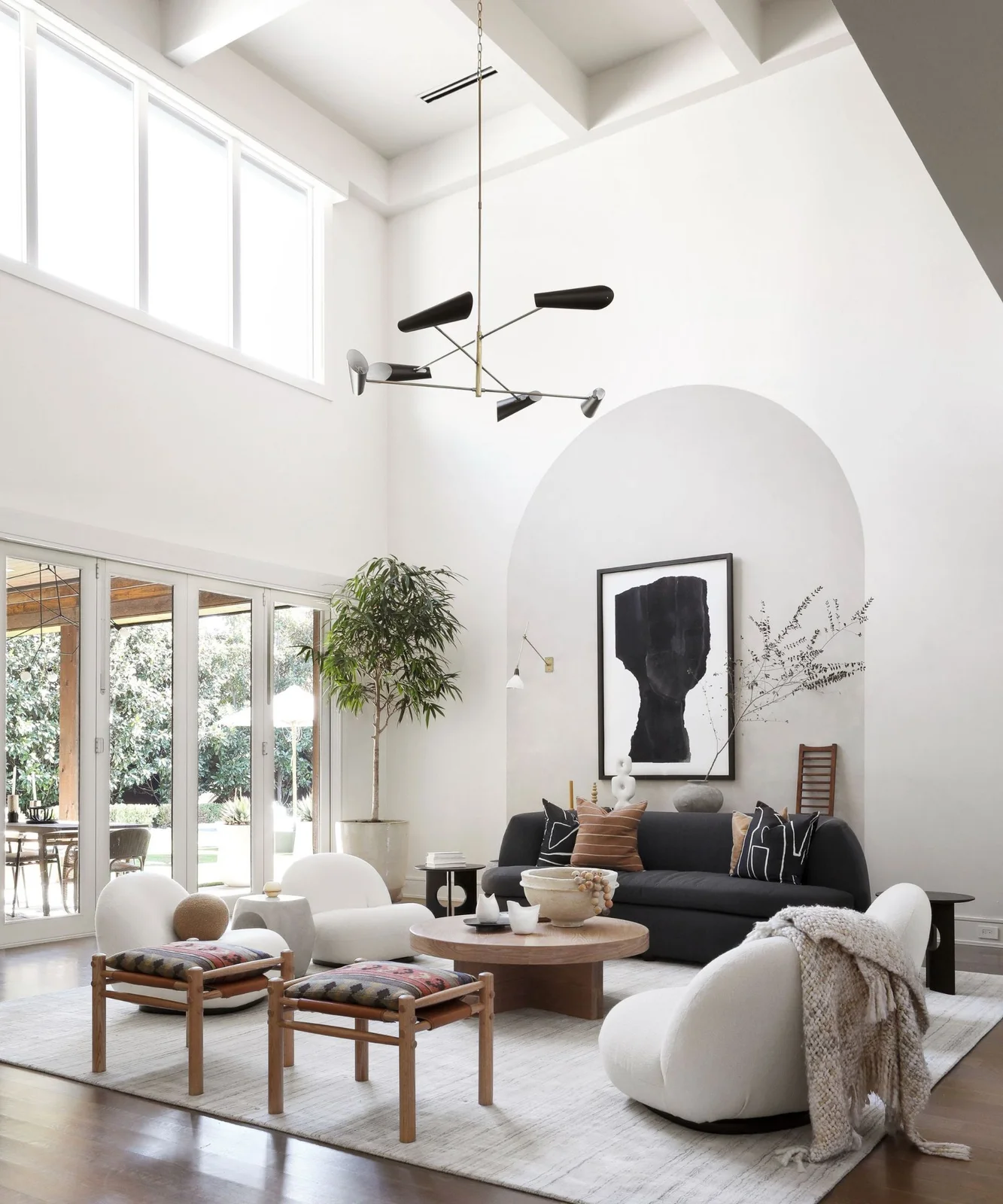 Rounded white chairs and timber stools face a dark sofa around a round timber table beneath a tall arched wall recess.
