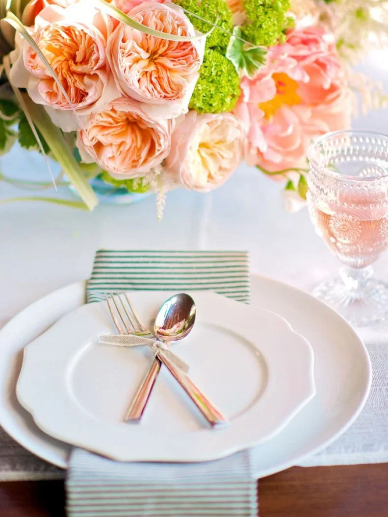 A spoon and fork tied together rest on a white scalloped plate above a green-striped napkin beside pale peach flowers