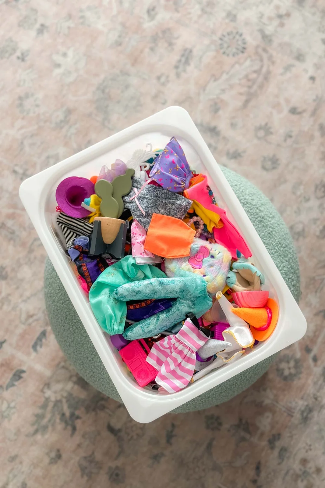 White open bin containing colorful fashion-doll clothes, miniature hats, bags and other accessories.