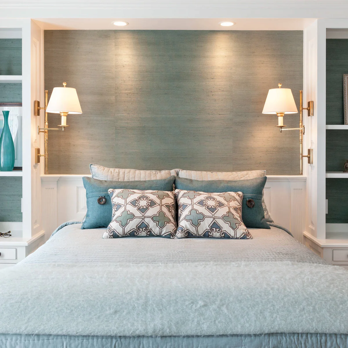 Two pale shaded warm metal wall lights are mounted on white piers beside a recessed bed with shelves and teal cushions.