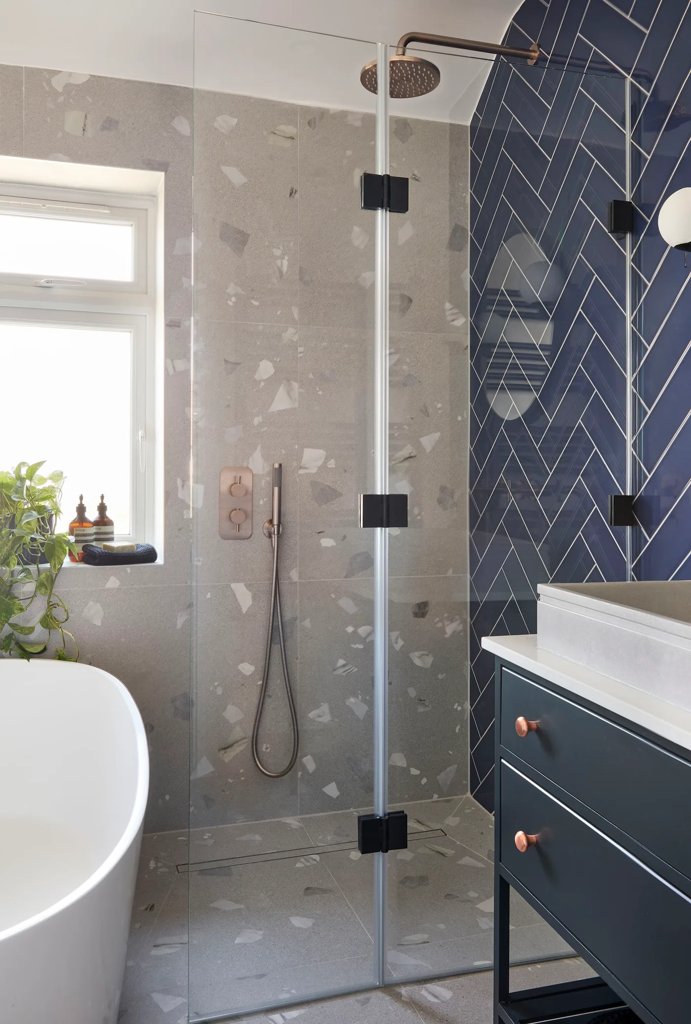 Navy herringbone tiles and a dark vanity sit beside a glass shower enclosure with pale speckled wall tiles