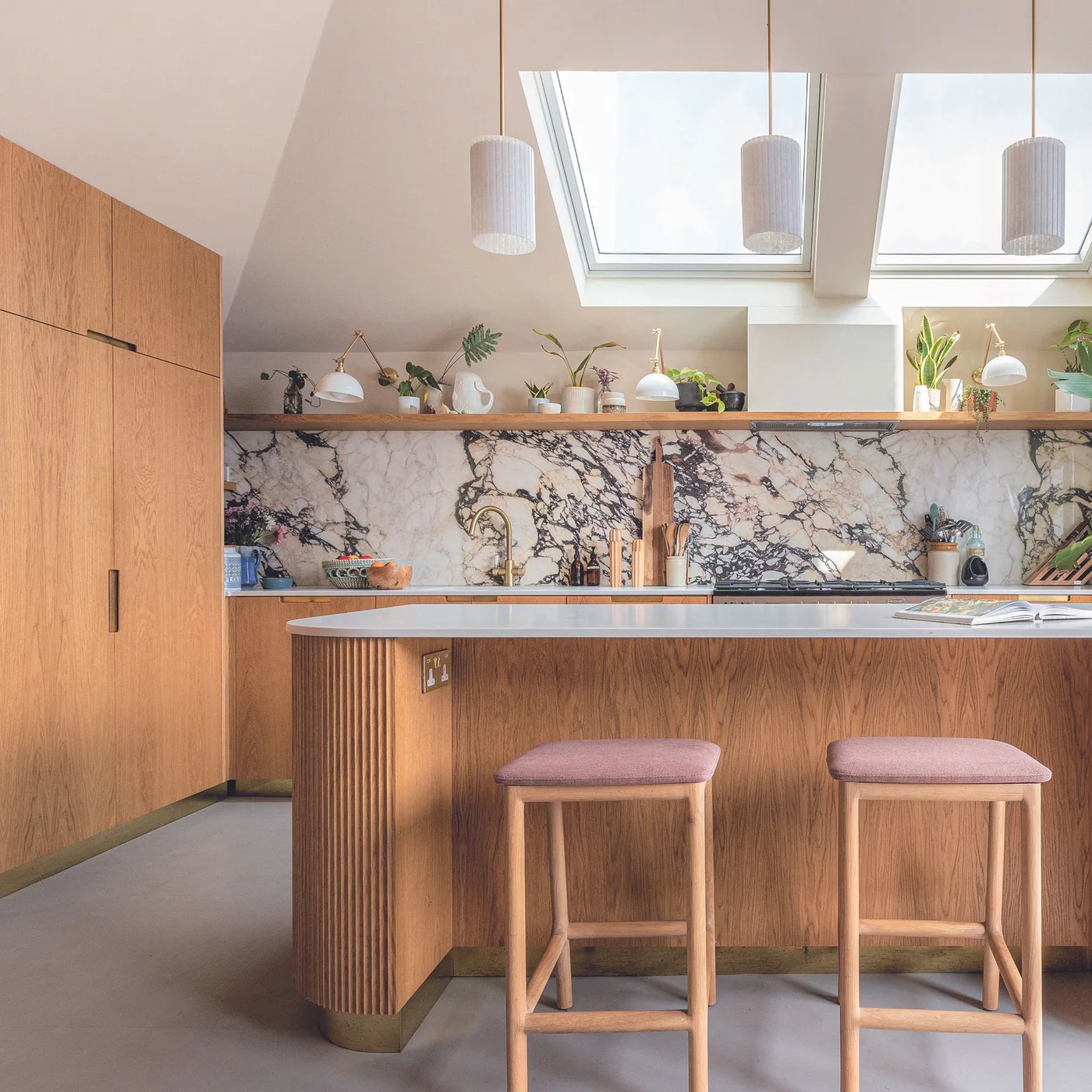 A ribbed wooden island and pink-seated stools sit below pale pendants beside flat wooden cupboards