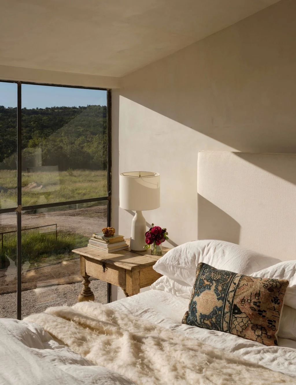 A pale bed with patterned cushions sits beside a simple wood bedside table and white shaded lamp near a tall dark-framed window beneath a sloping ceiling