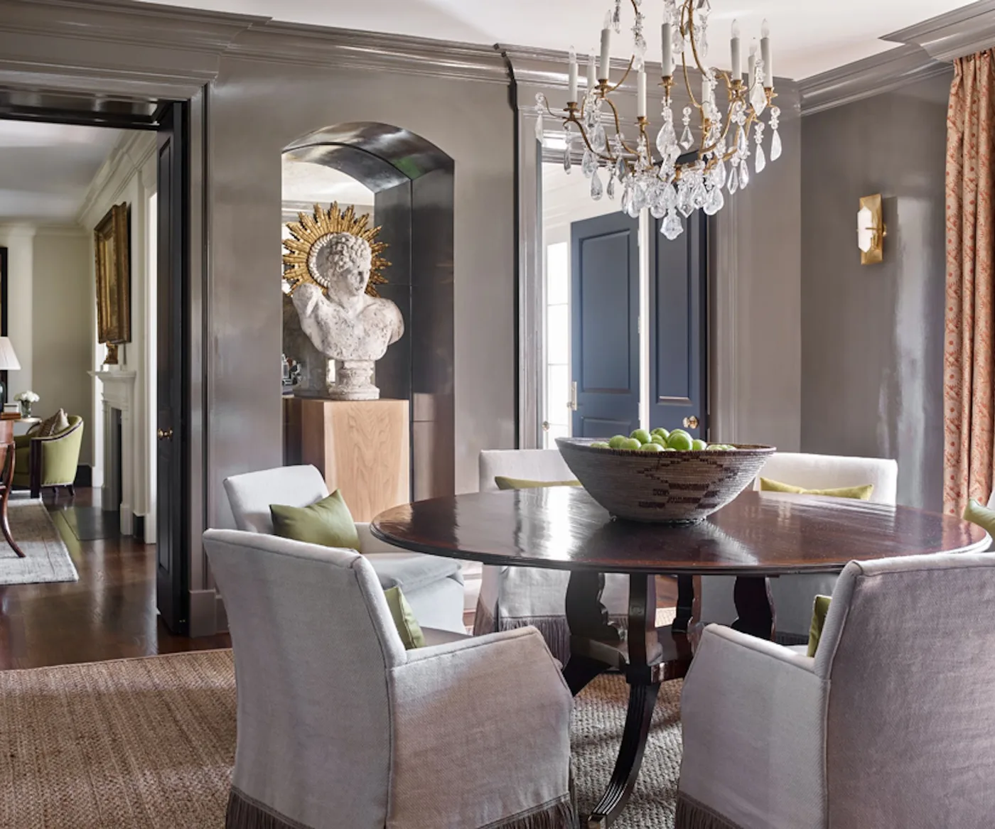 Gray dining room with a dark round table, pale upholstered armchairs, olive cushions, ornate chandelier and sculpture in an arched niche