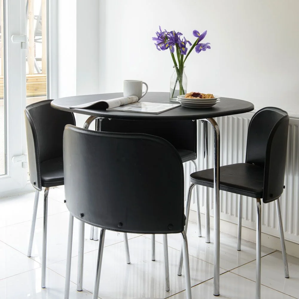 Three black curved-back chairs surround a small dark round table beside pale walls, a radiator and a glazed door.