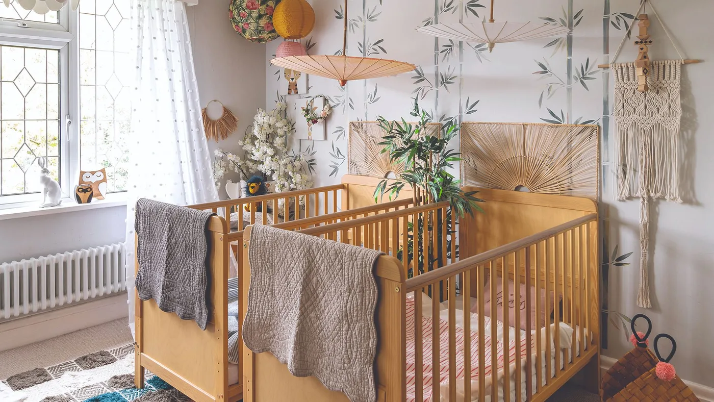 Two warm wooden cribs with gray throws against pale leafy wallpaper, woven panels and a fringed wall hanging.