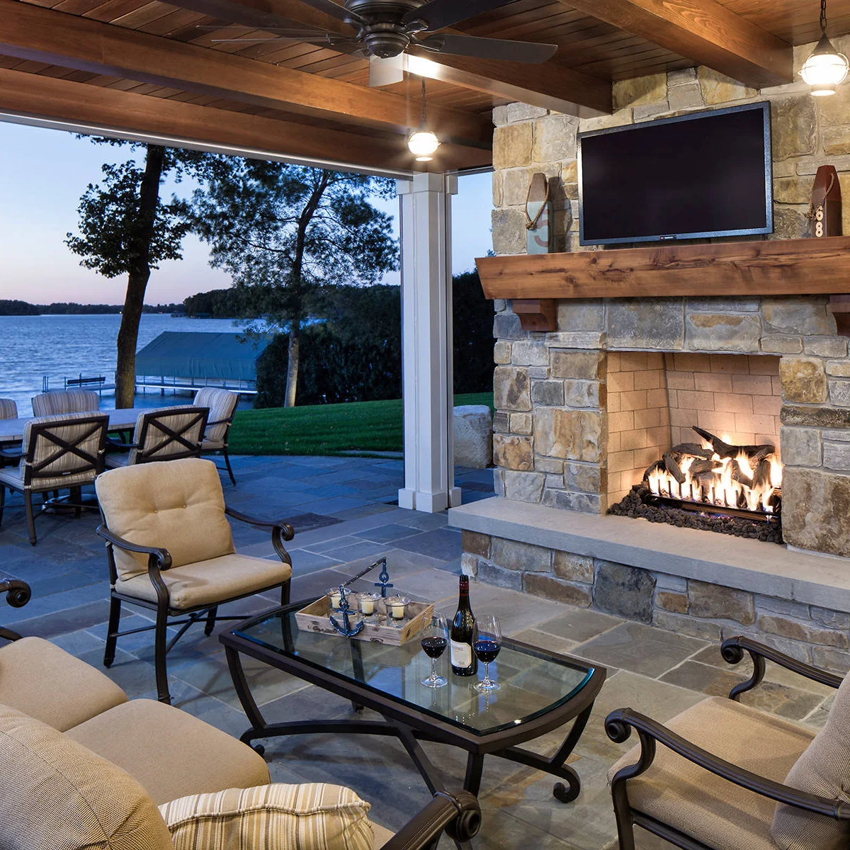 Pale cushioned chairs and a glass table sit beneath a timber patio ceiling beside a stone fireplace, with a lake visible outside.
