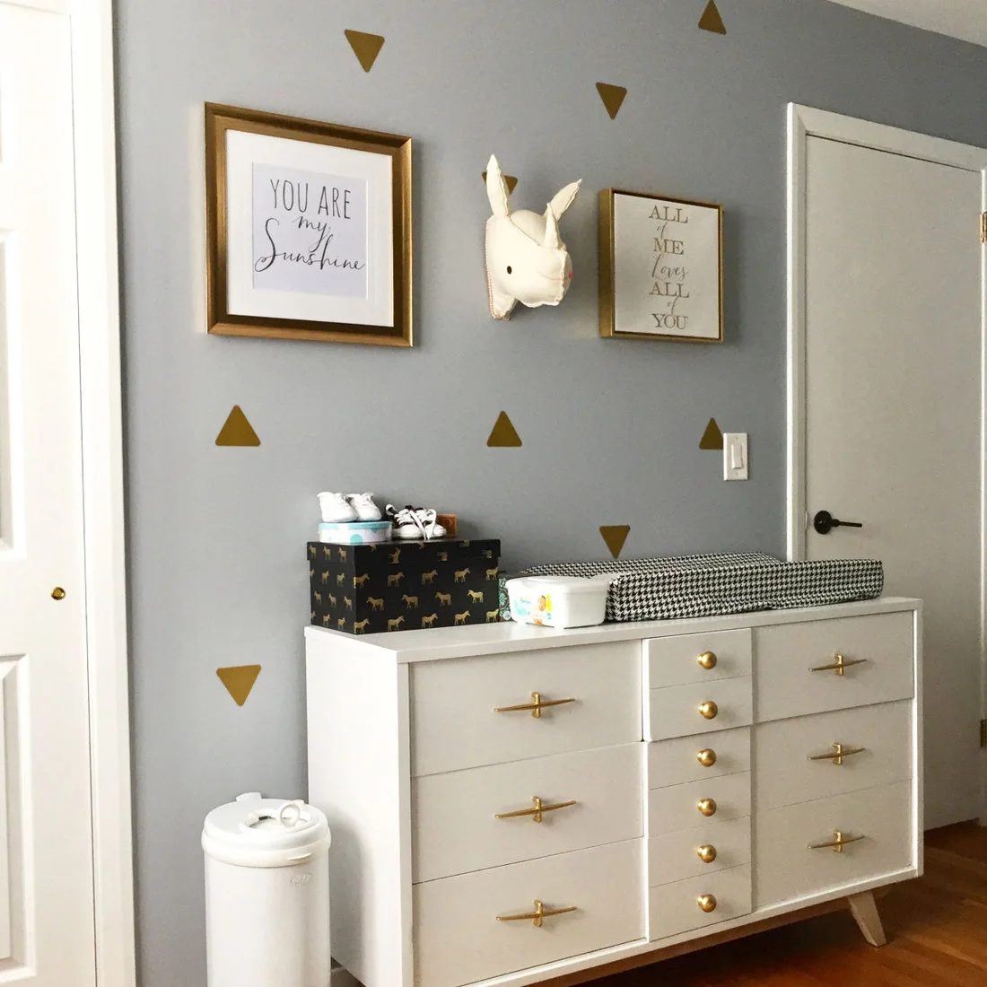 A low white changing dresser with long warm-metal handles stands beneath a gray wall with gold triangles beside a lidded floor bin.