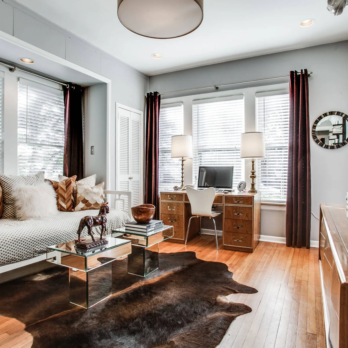 A white daybed faces two small mirrored tables in a home office with a wood desk and burgundy curtains.