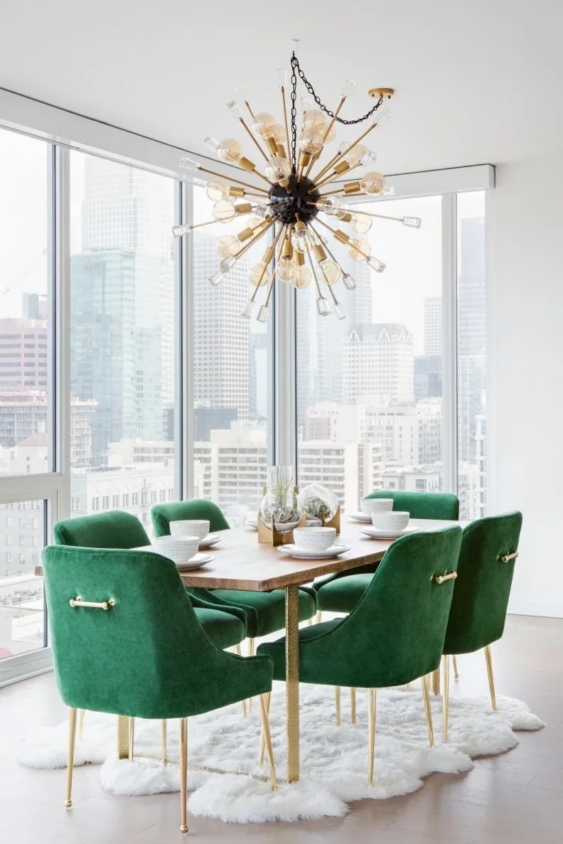 Radial chandelier with gold-toned arms, clear lamp surrounds, and a dark center above a wooden table and green upholstered chairs beside corner windows.