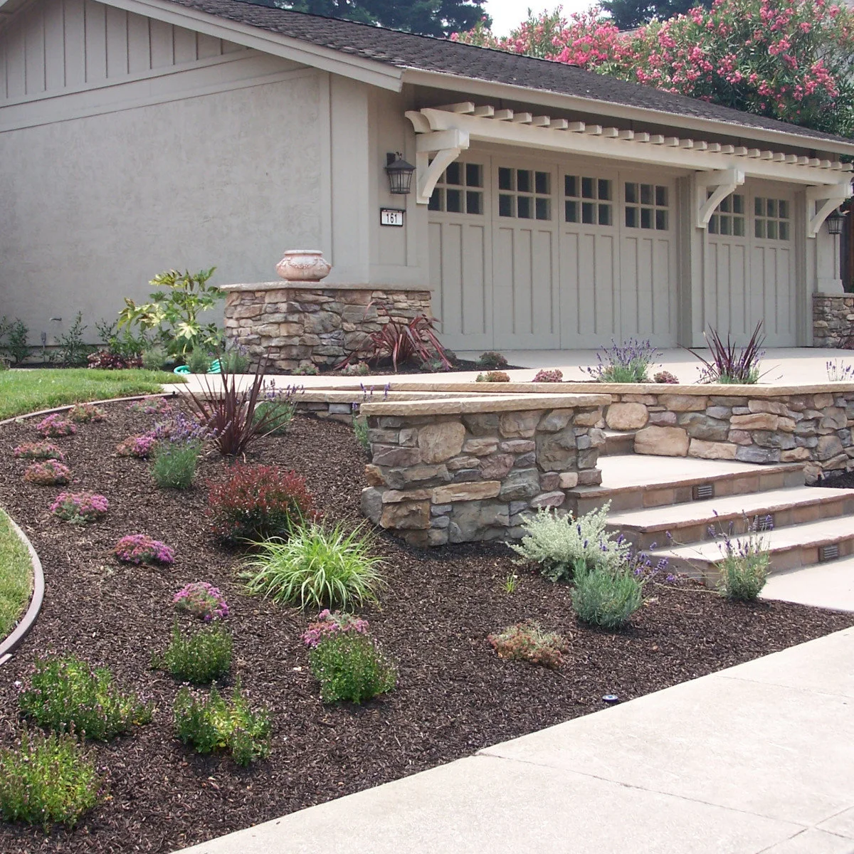 A broad mulched front bed with spaced grasses, low flowering plants, stone-faced steps and pale garage doors.