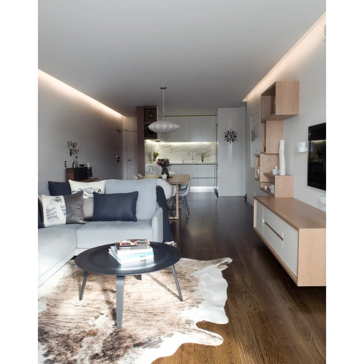 Long narrow room with a pale sectional, dark round coffee table, dining table and white kitchen beyond.