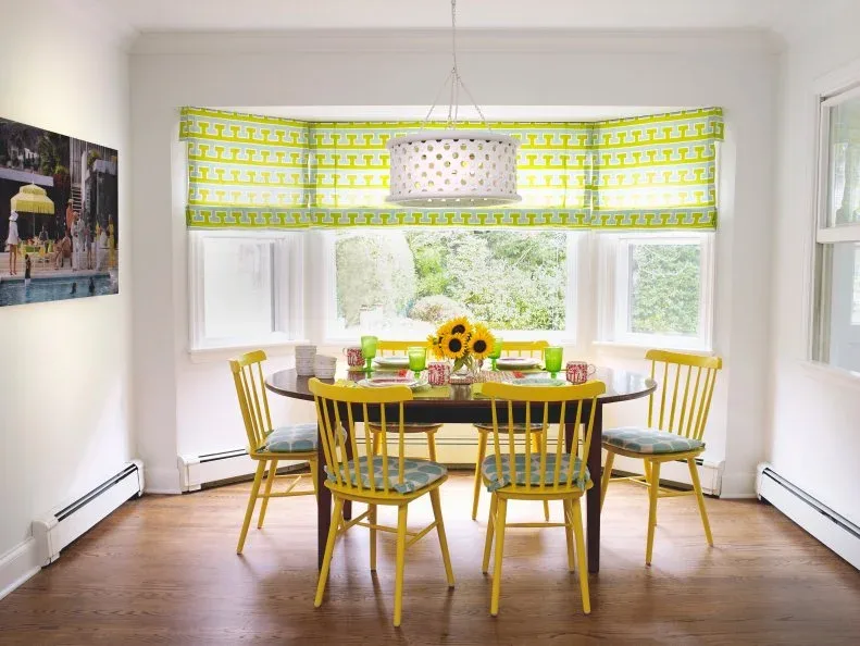 Bright dining nook with yellow spindle chairs, an oval dark table, green-and-white patterned window fabric and a light drum pendant
