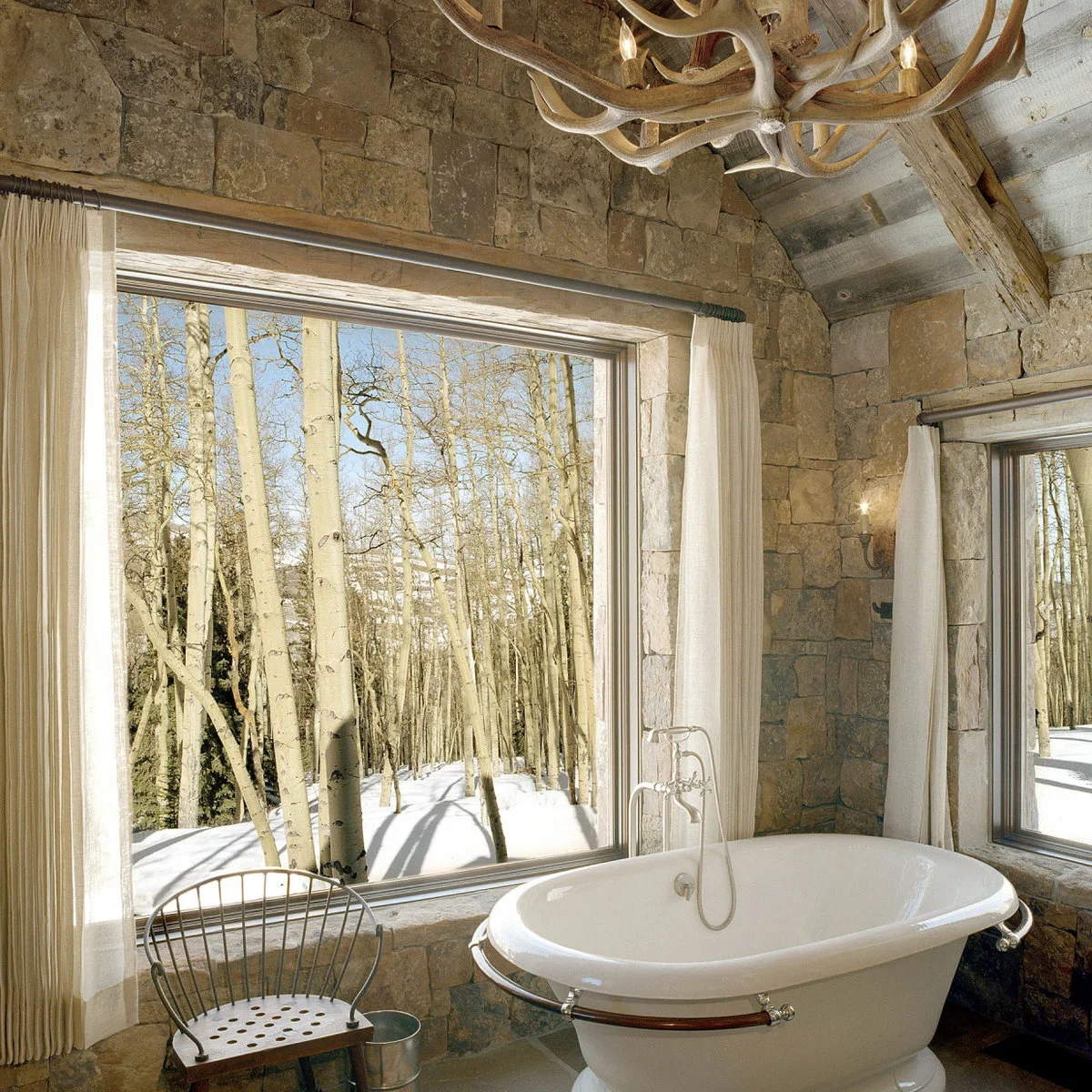 Plain pale curtains frame a broad window beside a white bath in a bathroom with rough stone walls and timber beams.