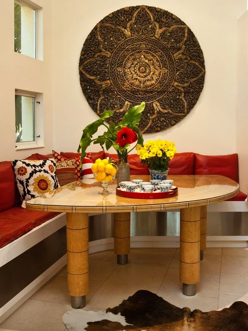 Red-cushioned corner banquette with patterned pillows and a white base, rounded pale wooden table on cylindrical legs, large circular wall ornament, flowers, and small windows.