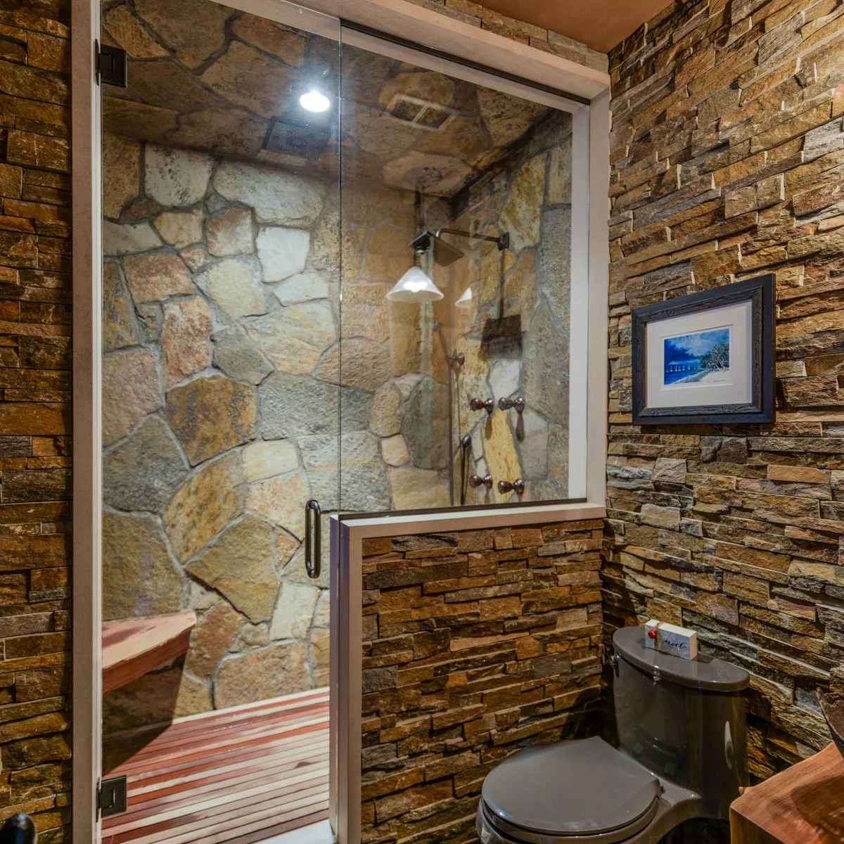 A shower with large irregular stone-textured walls behind glass, beside finely layered brown cladding and a dark toilet.