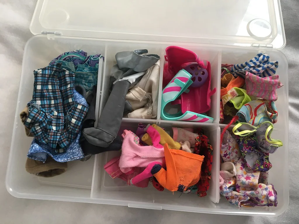 Open clear lidded box with divided compartments holding doll clothes, swimwear and small accessories.