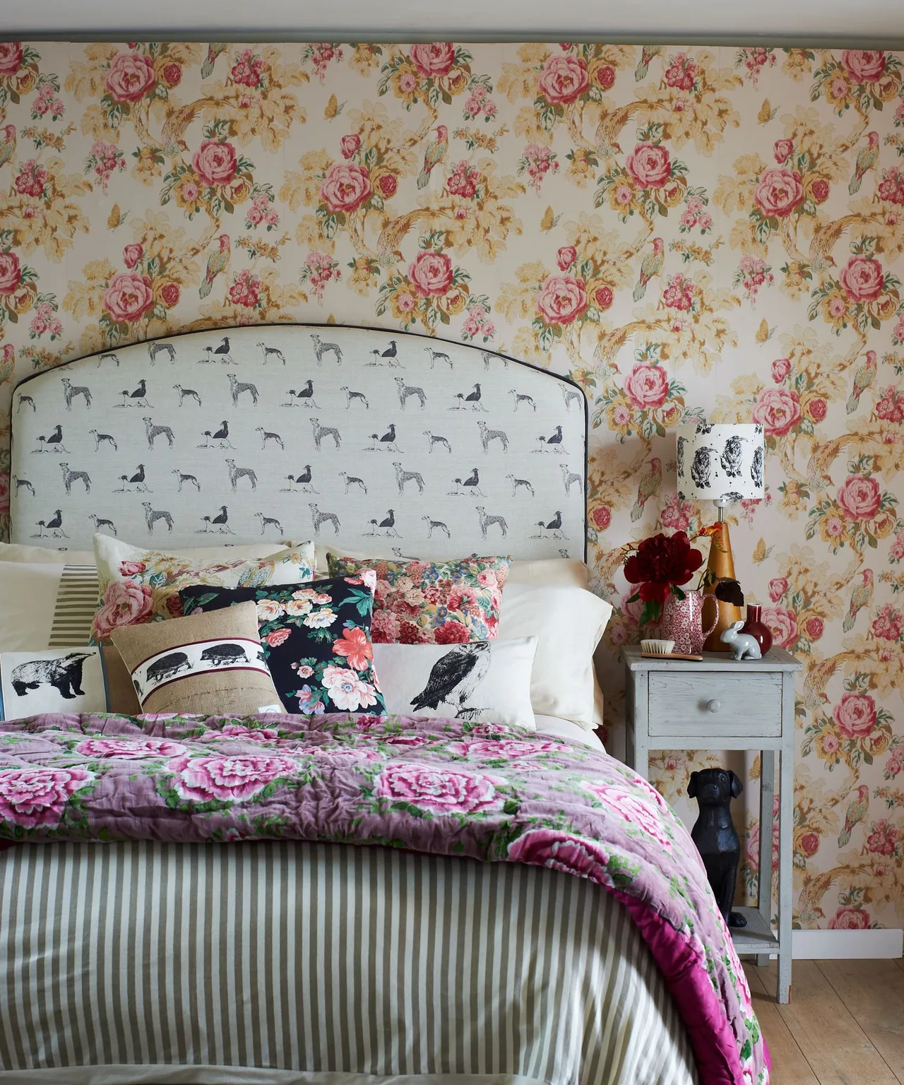 Bedroom with rose wallpaper, pale animal-patterned curved headboard, mixed cushions, floral bedcover and striped bed skirt.
