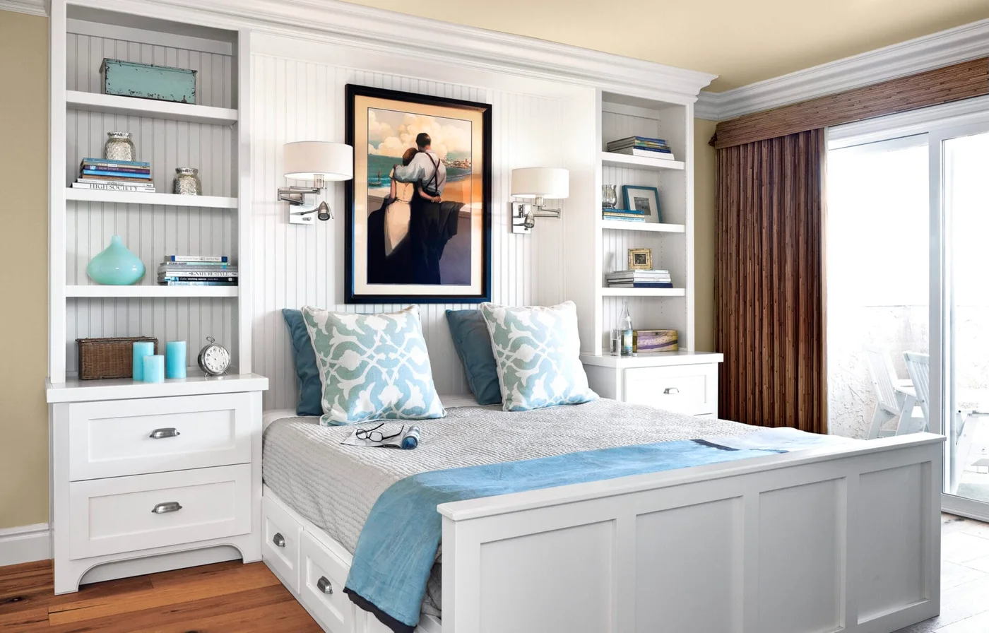 A white guest bed is framed by beadboard-backed shelves, bedside drawers and wall lamps, with blue cushions and a blue throw.