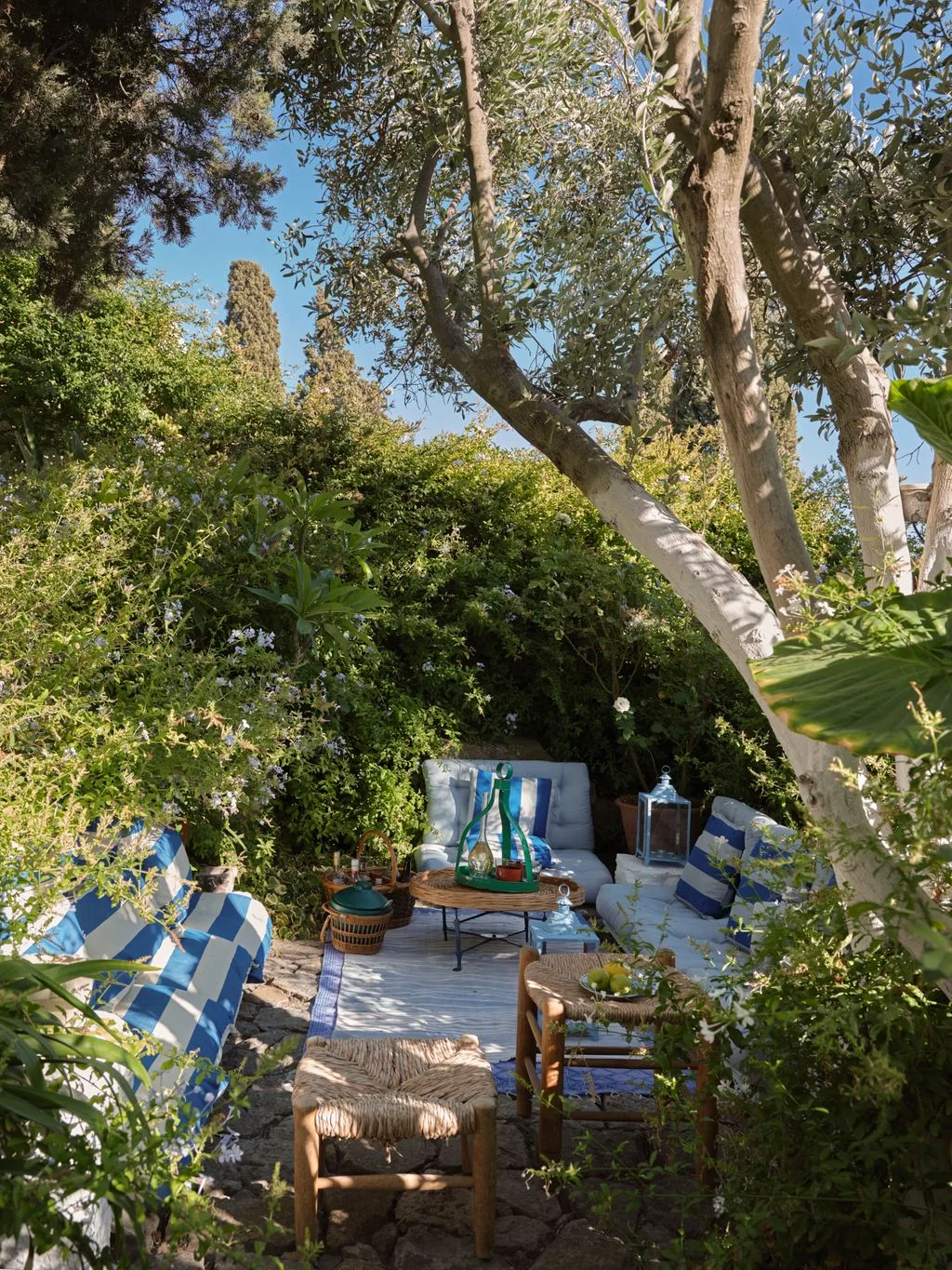 Sheltered garden seating with blue-and-white striped cushions, woven stools and small tables among greenery