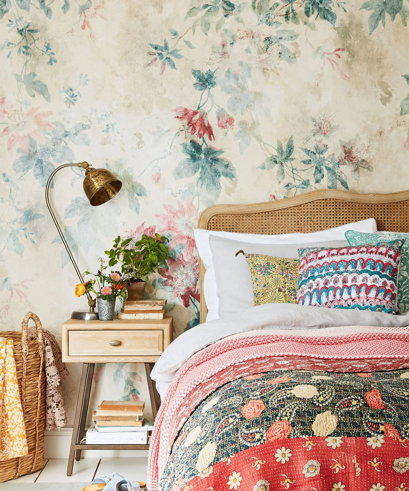 Bedroom with pale multicolored floral wall, curved woven headboard, patterned pillows and bedcover, wooden nightstand and curved reading lamp.