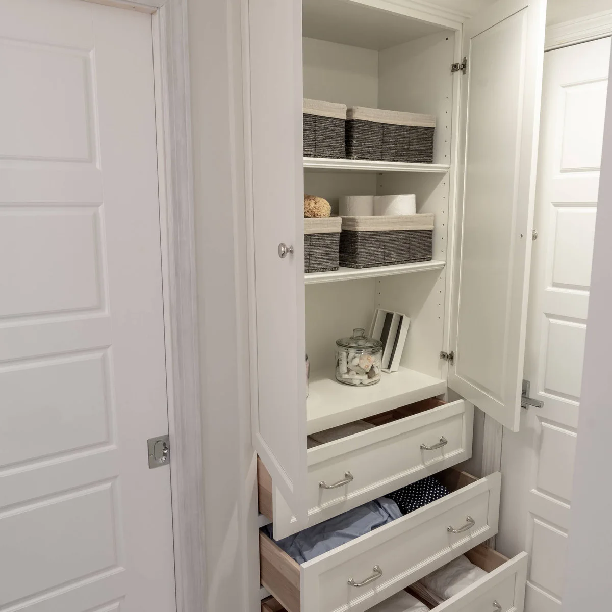 Narrow white cabinet with lined baskets on shelves and open lower drawers containing pale and patterned linens