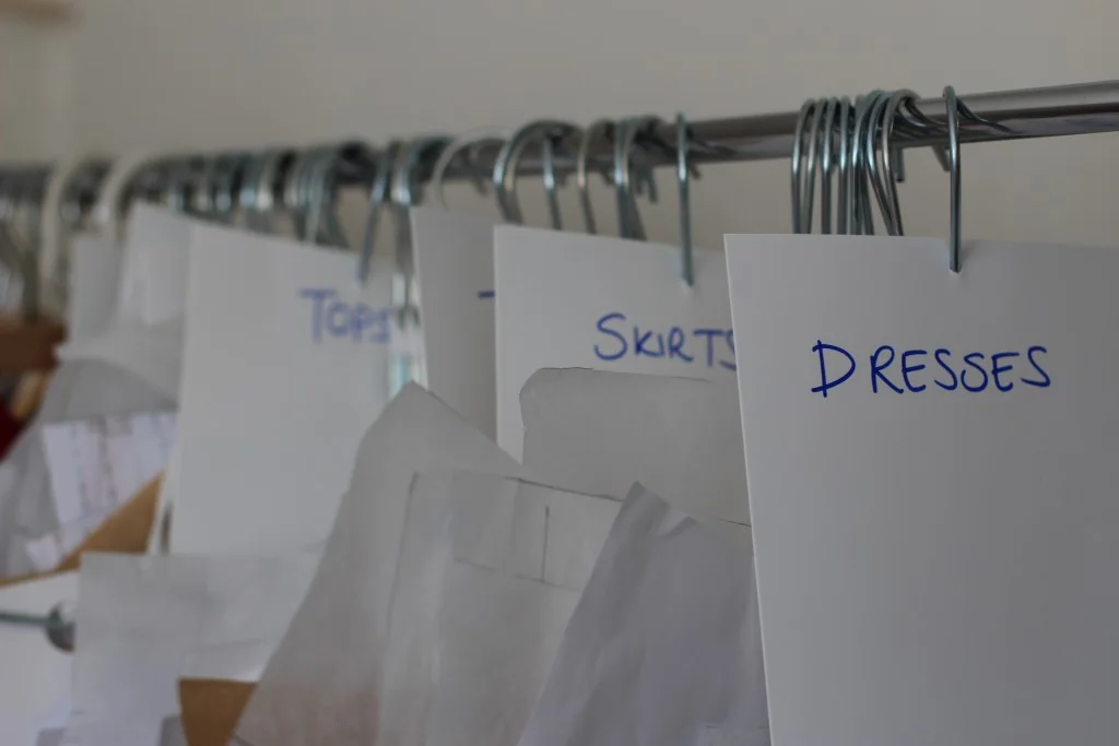 Paper sewing patterns hang from metal hooks on a rail, with handwritten category cards separating tops, skirts and dresses.