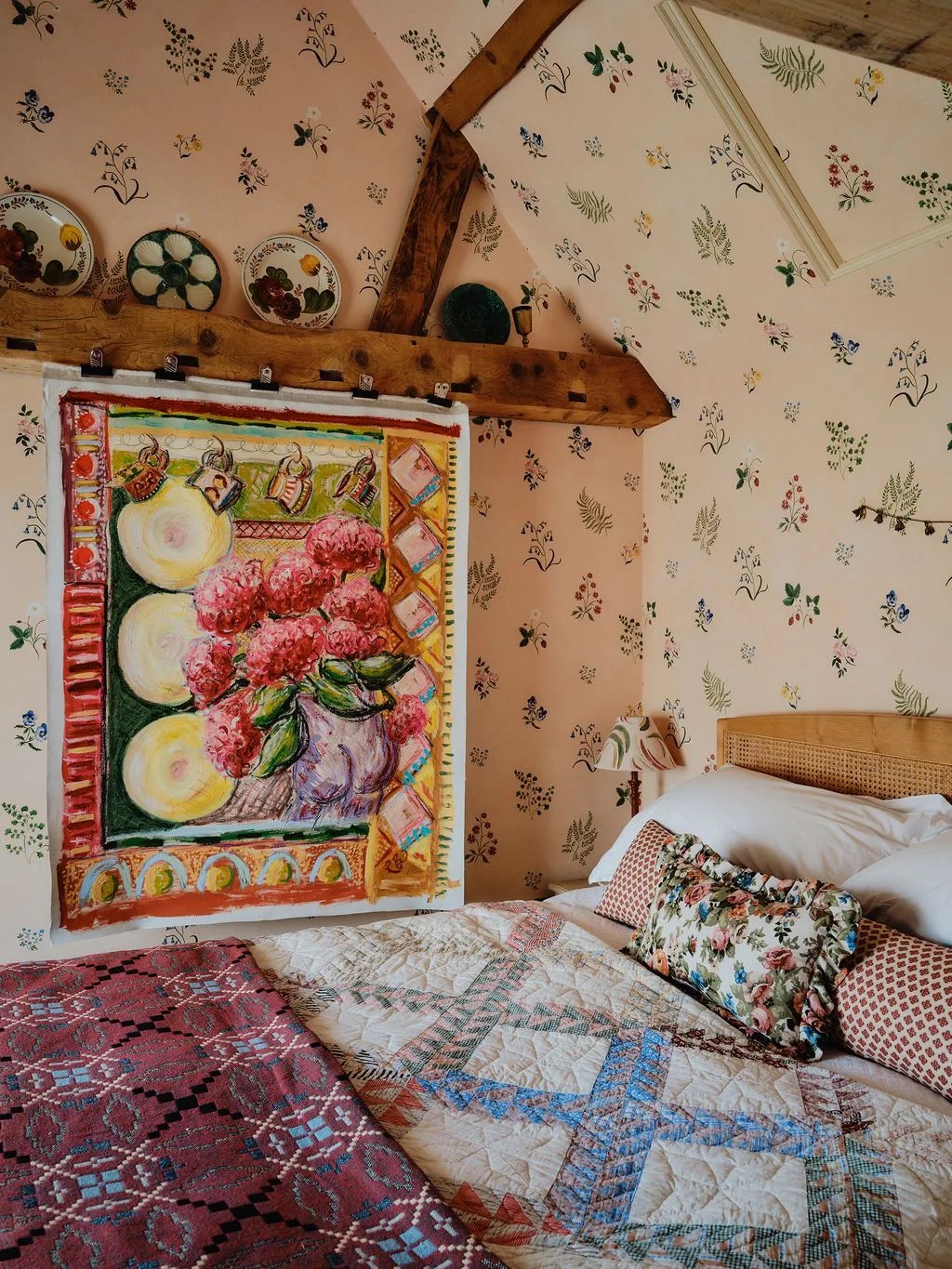 Attic bedroom with pale floral wallpaper, exposed beam, large colorful wall hanging, patchwork bedding and decorative plates