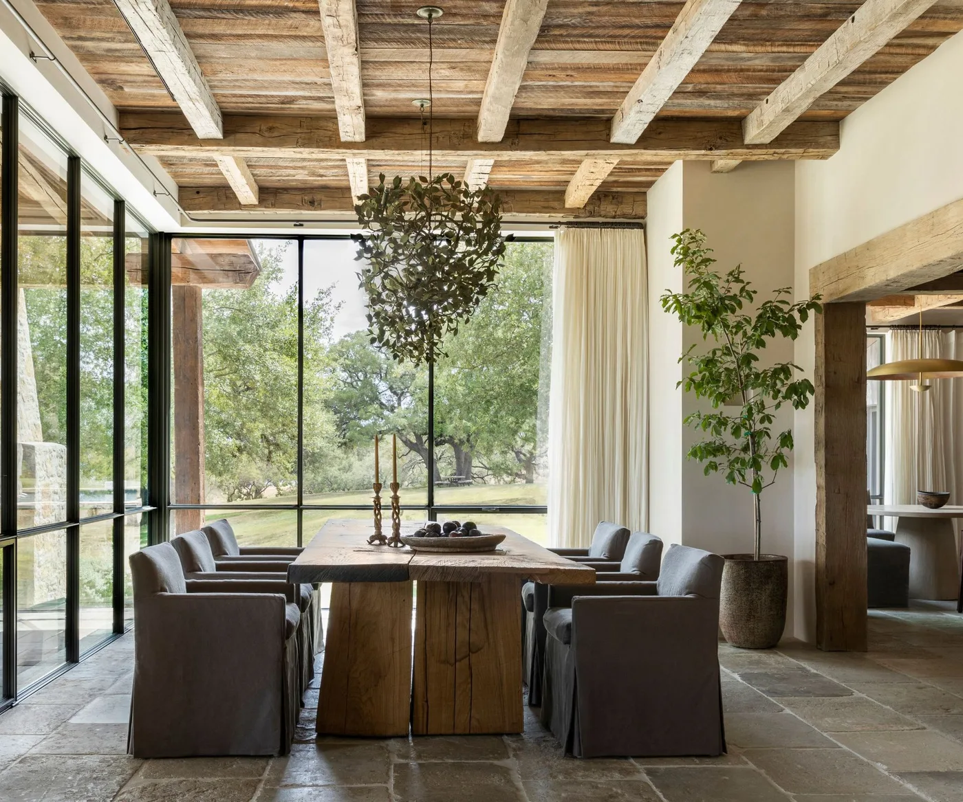 Gray skirted chairs surround a large wooden dining table beneath wooden ceiling beams and a leafy hanging fixture beside tall windows