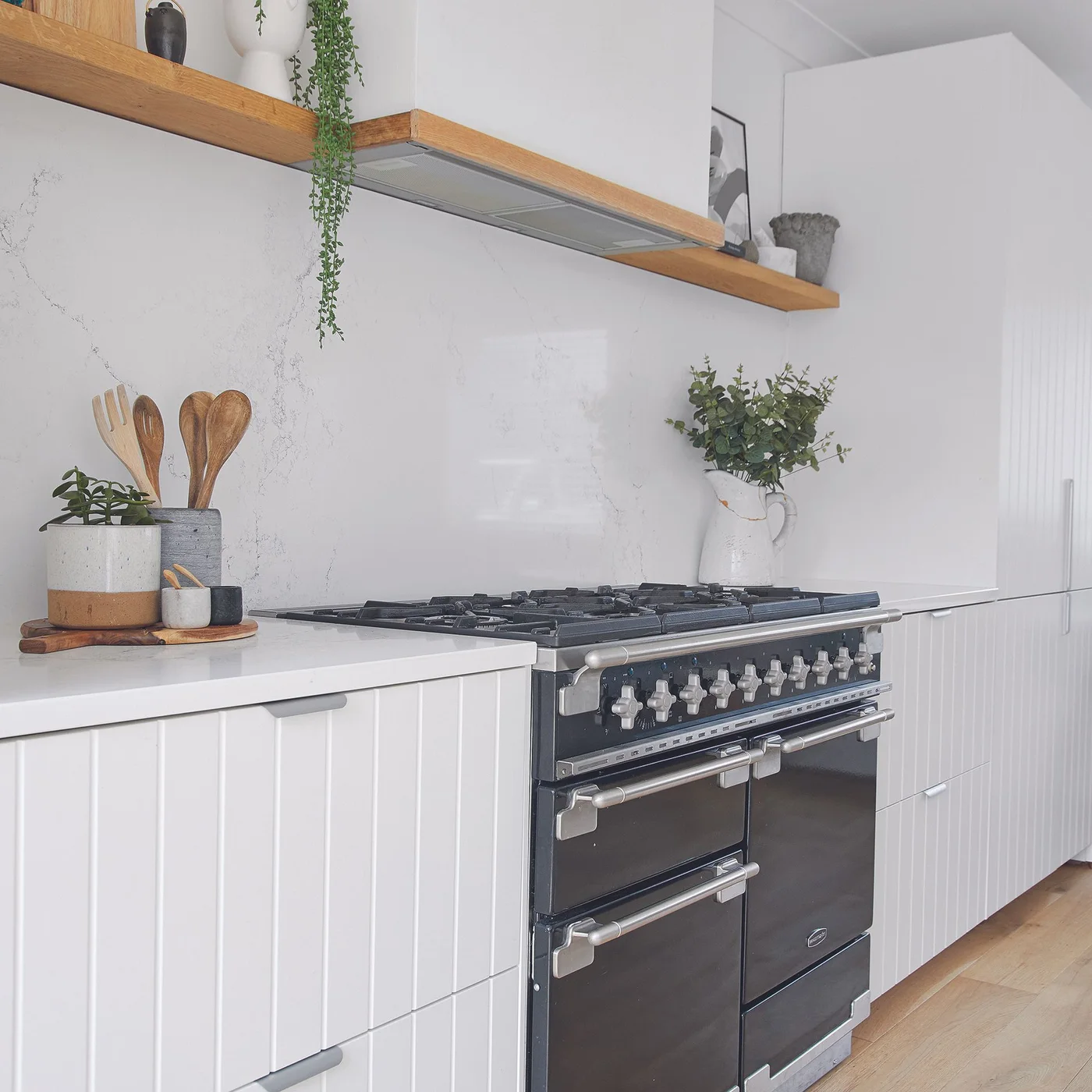 White vertically grooved cabinet fronts surround a black range beneath a plain white hood with timber trim.