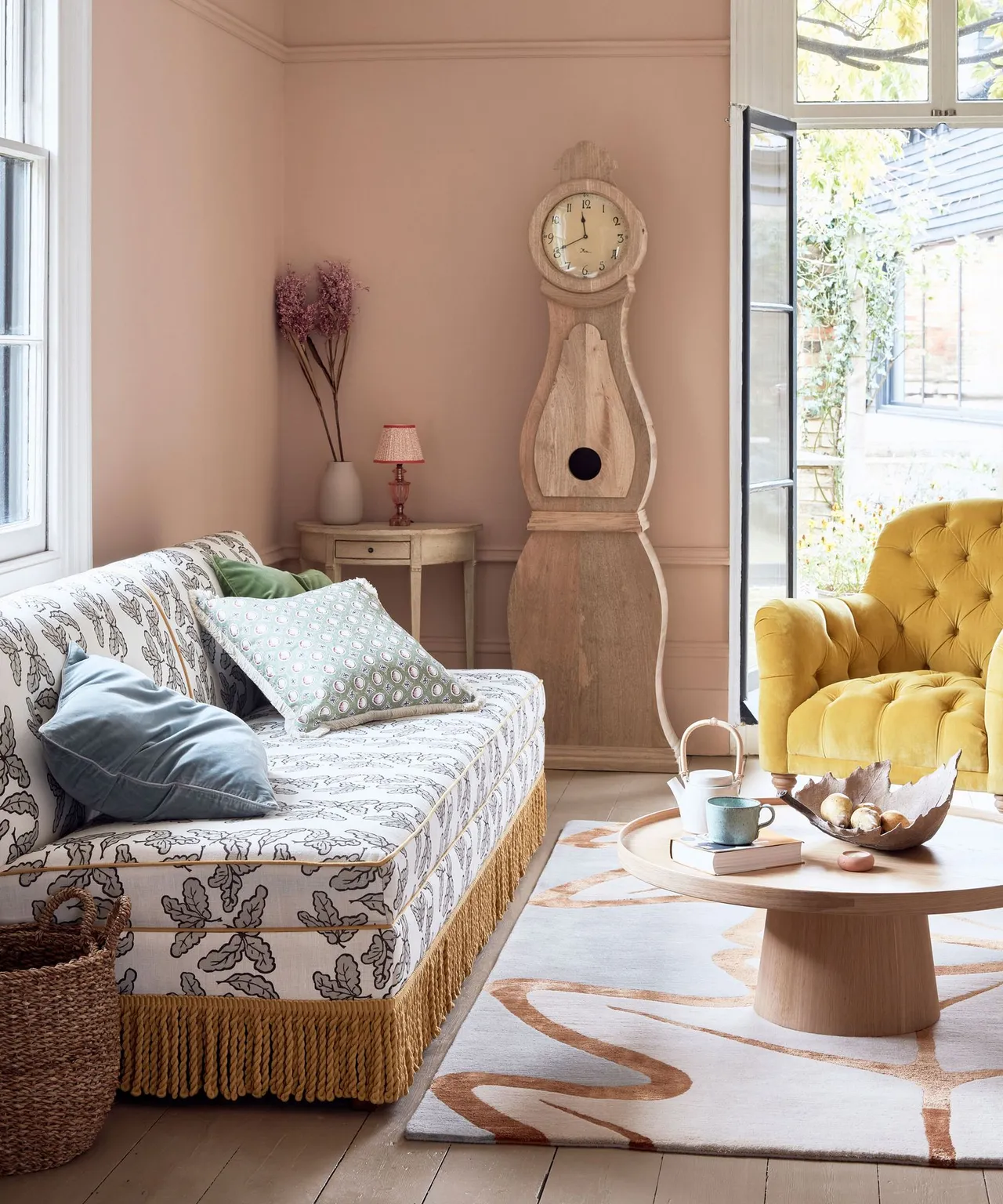 Living room with patterned fringed sofa, yellow buttoned armchair, pale tall clock, small bedside-style table, pink walls and round wooden coffee table.