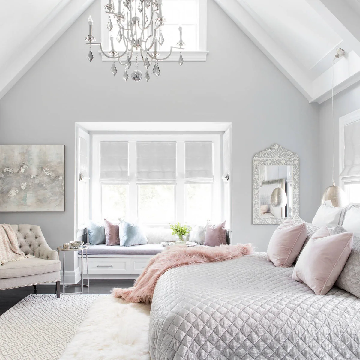 Bedroom with a pale gray bedcover, blush pillows and throw, and a window seat with pink and blue cushions