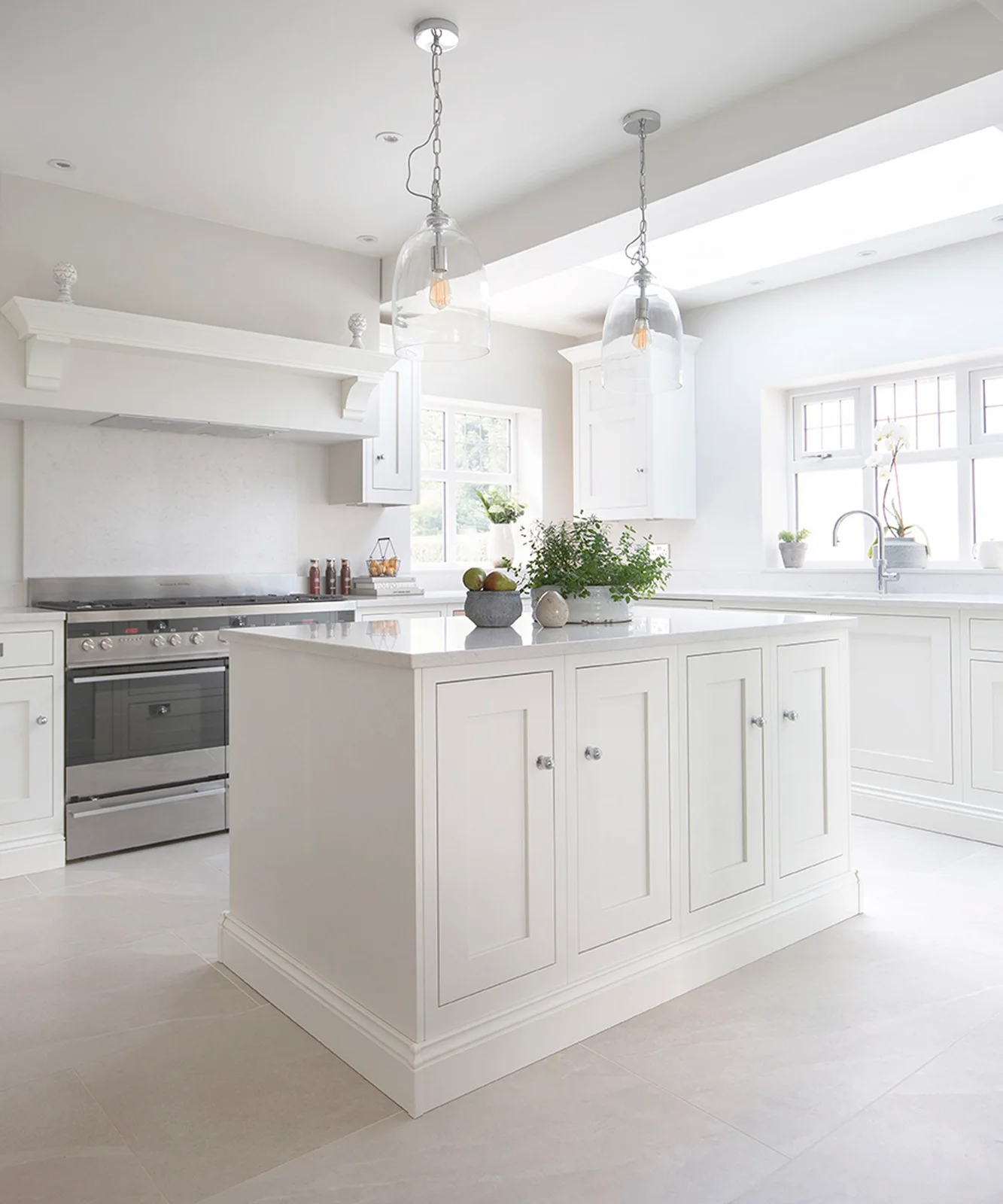 Soft-white framed kitchen cabinets and a matching island sit beneath glass pendants beside a pale floor and stainless range.