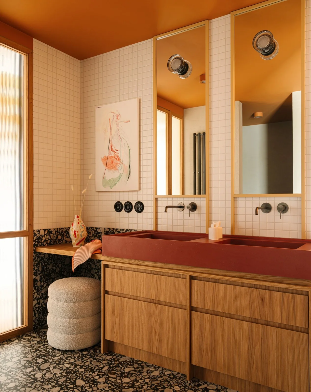 A red double bathroom basin sits on a wood vanity beneath tall framed mirrors beside white square tiles, an orange ceiling and a rounded upholstered stool