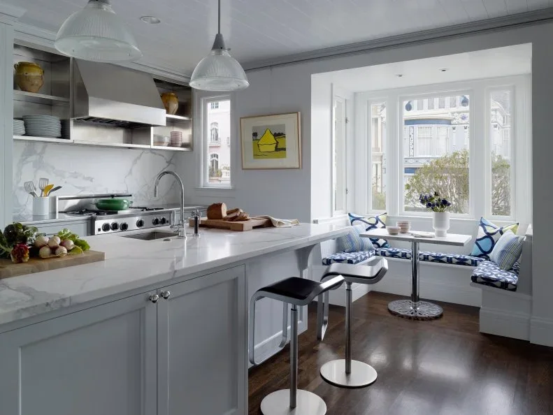 A gray paneled island with two streamlined stools stands beside open kitchen shelving and a window breakfast seat with blue patterned cushions