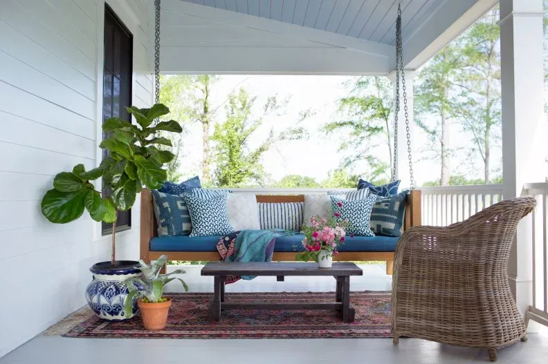 White country porch with wood swing, blue cushions, red patterned rug and woven armchair