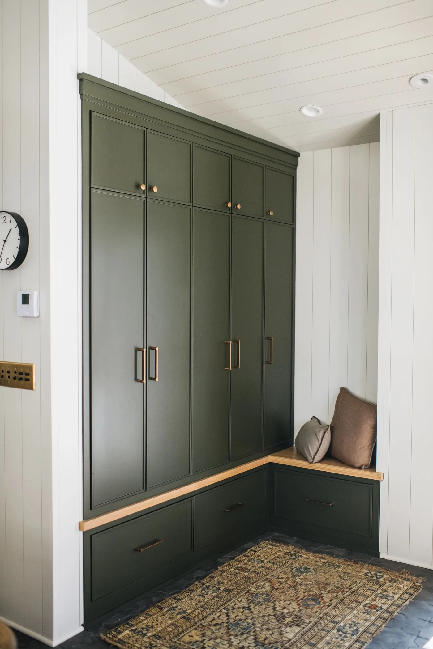 Green mudroom cupboard with L-shaped wooden bench top, lower drawers, white paneled walls and patterned rug.