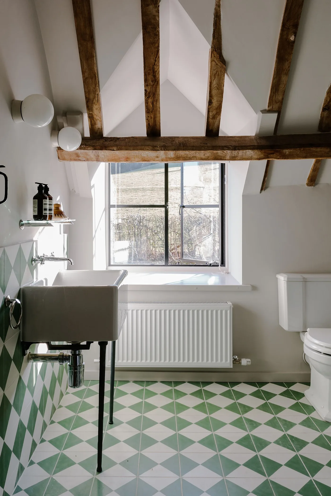 Bathroom with exposed wooden beams, green and white geometric tiles, a wall basin on dark legs and a bath beneath the window.