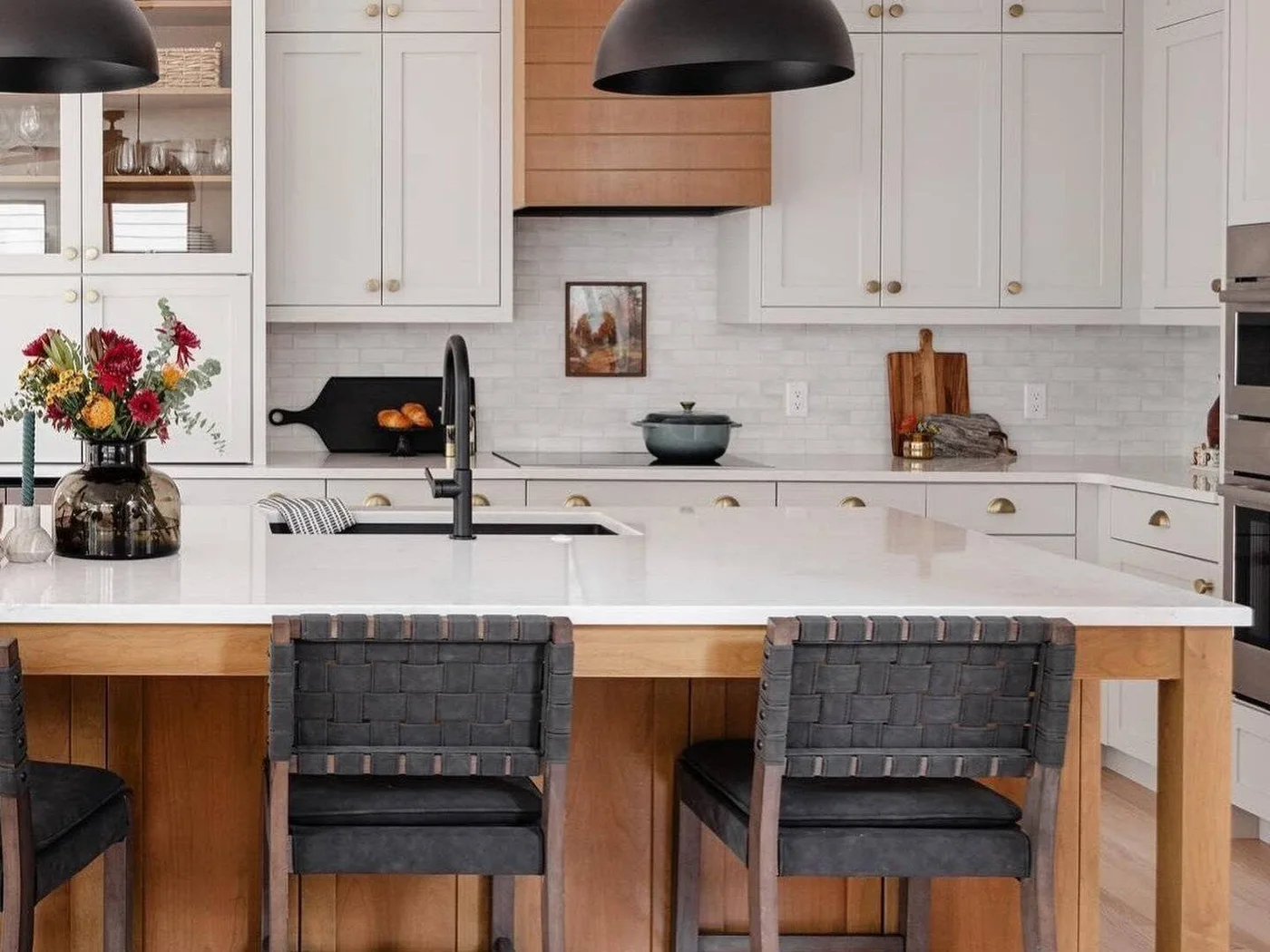 White rectangular backsplash tiles sit beneath white kitchen cupboards beside a timber hood and dark island stools.