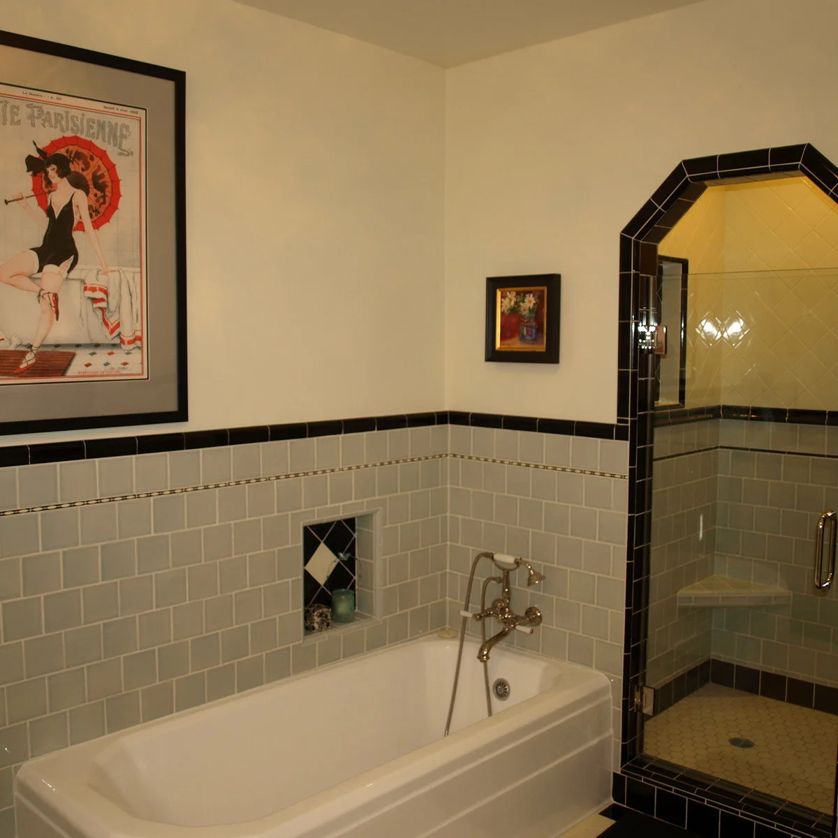 Dark trim outlines an angular shower opening beside gray rectangular wall tile, a white bath and framed wall art.