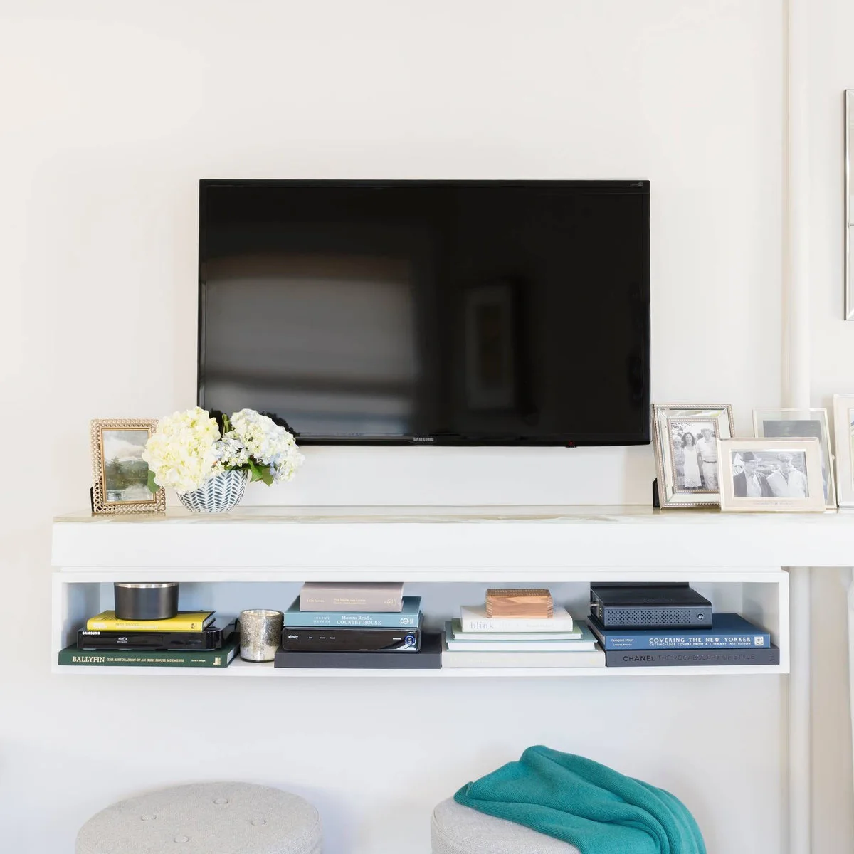 White wall-mounted media shelf below a television with books, framed photographs and two small stools