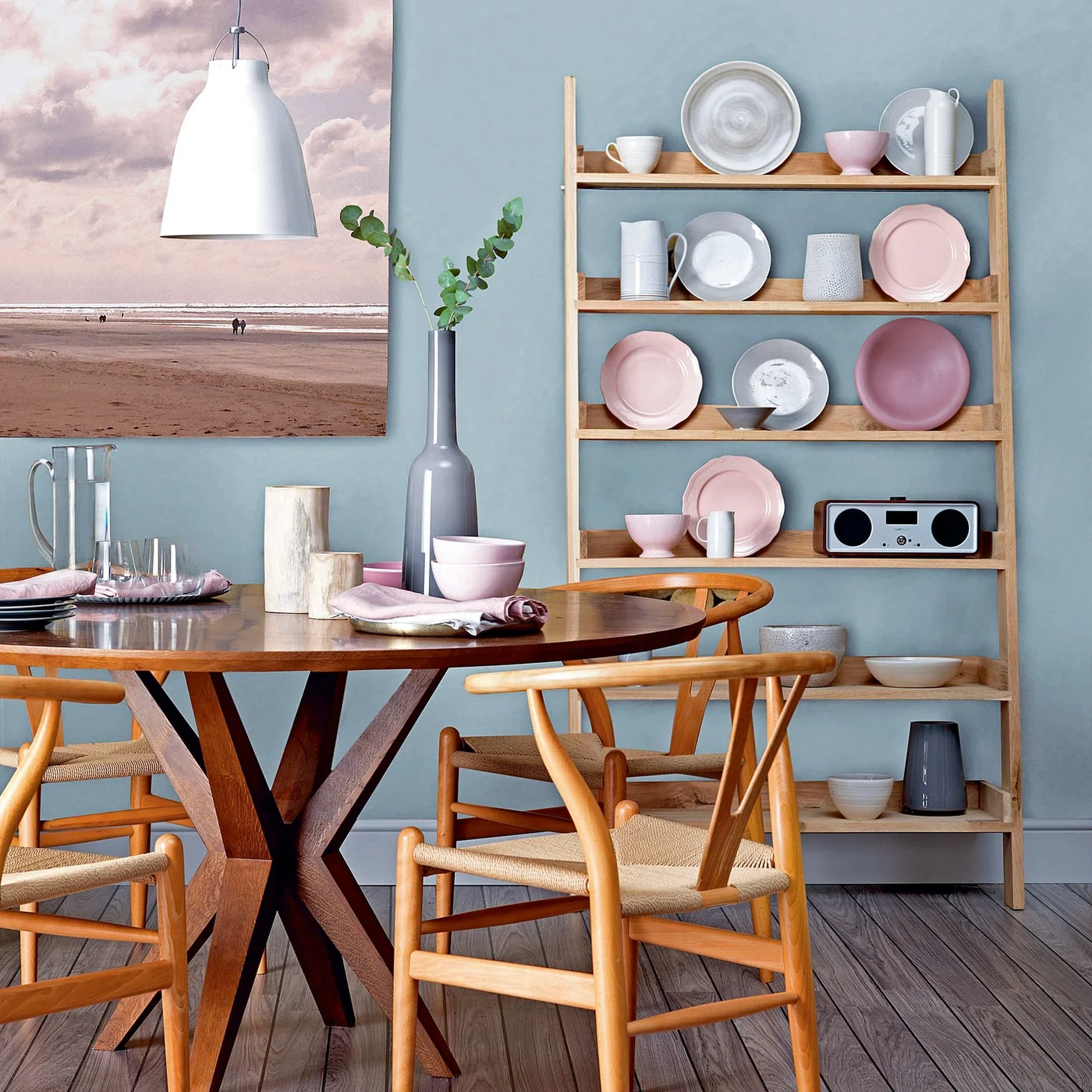 A slim wood wall rack holds pink and pale plates, cups and a small radio beside a round wood dining table and woven-seat chairs.