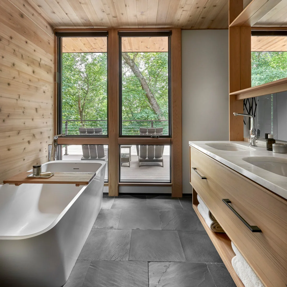 A white freestanding tub beside a timber wall and wood vanity facing tall windows