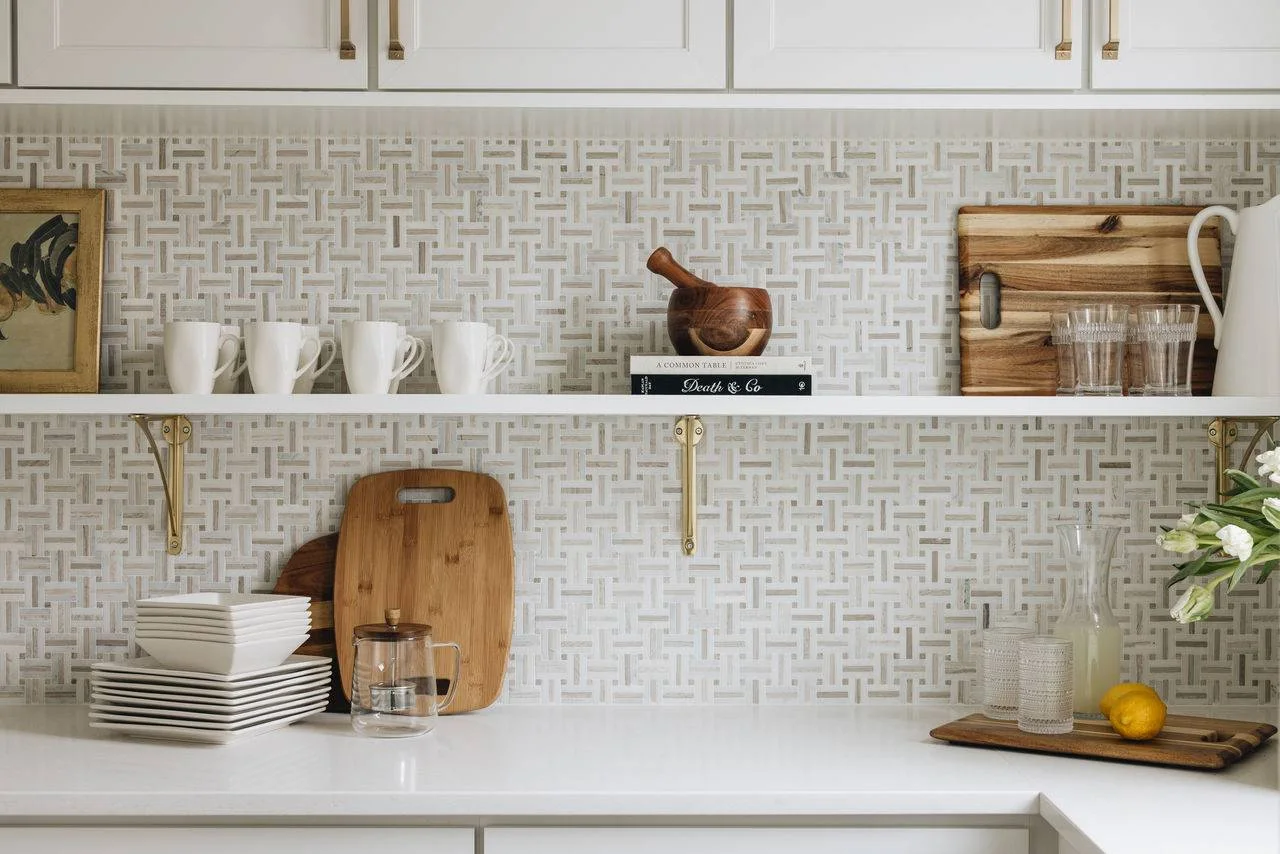 A white-and-beige basketweave mosaic fills a kitchen backsplash behind a slim shelf and white dishes.