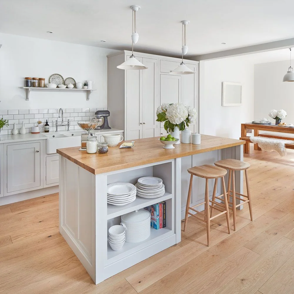 A pale kitchen island with a wooden top and stools stands beside a wooden dining bench and white cabinets