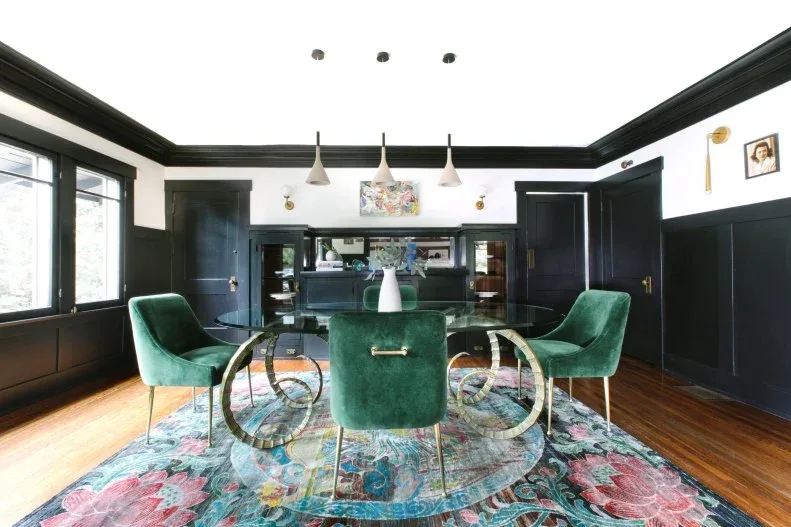 Dining room with dark lower walls and trim, green upholstered chairs, a glass table, three white pendants and a colorful rug